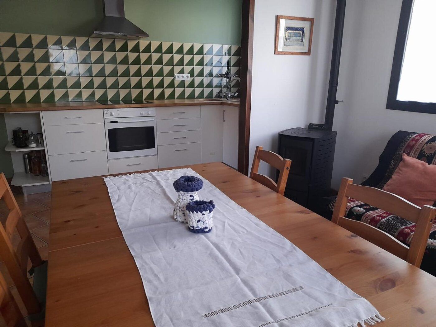 Ein Essbereich mit einem Holztisch, Stühlen, einer weißen Tischdecke und einer Küche im Hintergrund mit einer grün-weißen Fliesenwand.