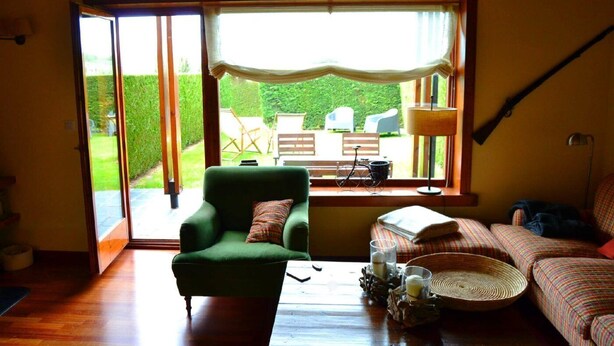 A cozy living room with a green armchair, a sofa, and a dining table with a lamp and glasses. A large window offers a view of a garden and outdoor furniture.