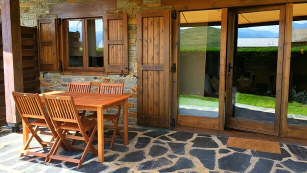 A stone patio with a wooden table and chairs, stone walls, and a view of a grassy area through glass doors. - Ground Floor 3 rooms Pool, Fireplace, Private Garden & Views (Olopte)