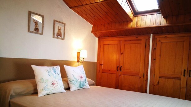 A bedroom with a wooden ceiling, a bed with two pillows, a nightstand, and a wall-mounted lamp.