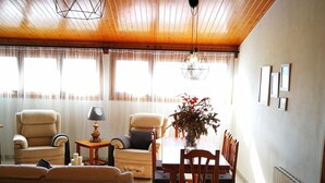 A cozy living room with a wooden ceiling, a dining table with chairs, a sofa, a lamp, and a vase with flowers.