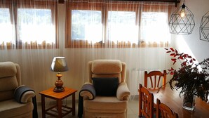 A cozy living room with a wooden ceiling, a beige armchair with a cushion, a small wooden table with a lamp, and a dining table with chairs.