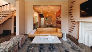 A spacious living room with a leather sofa, a coffee table, and a fireplace. There is a staircase on the left and a dining area in the background. - Bellwether House Summ (Louisville)