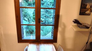 Uma área de refeições com mesa de madeira, cadeiras brancas, janela com vista para vegetação e um armário com cesta.