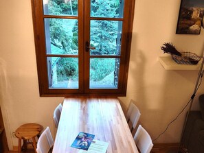 A dining area with a wooden table, white chairs, a window with a view of greenery, and a shelf with a basket. - Studio 2 rooms Pool, Cadí Views, Gym and Games Area (Enveitg)