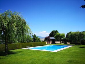 Una piscina rectangular con un cielo azul despejado al fondo, rodeada de césped cuidado y diversos árboles.