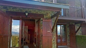 Un bâtiment rustique, doté de portes en bois et d’une façade en pierre, avec une véranda couverte et une vue sur la campagne.