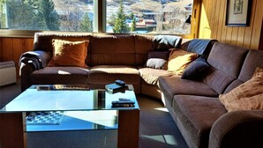 A cozy living room with a brown leather sofa, a glass coffee table, and a view of a snowy landscape through large windows. - Duplex 3 Bed Parking, Ski Storage & Games Room (La Molina)