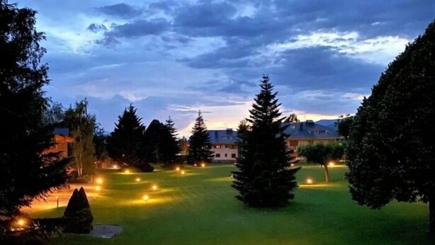 Campo de golf al anochecer con hoyos iluminados, rodeado de árboles y edificios.