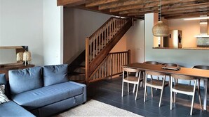 A modern living room with a blue sofa, wooden dining table, and chairs. A wooden staircase leads to an upper level.