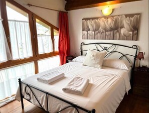 A bedroom with a bed, a window with curtains, a wall-mounted light, and a painting.