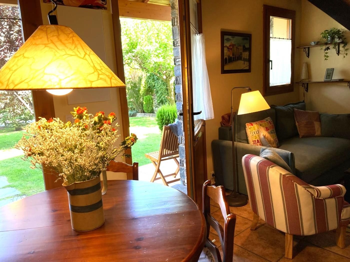 Una sala de estar acogedora con una mesa de madera, un sillón a rayas y un sofá. Se aprecia una ventana con vistas a un jardín.