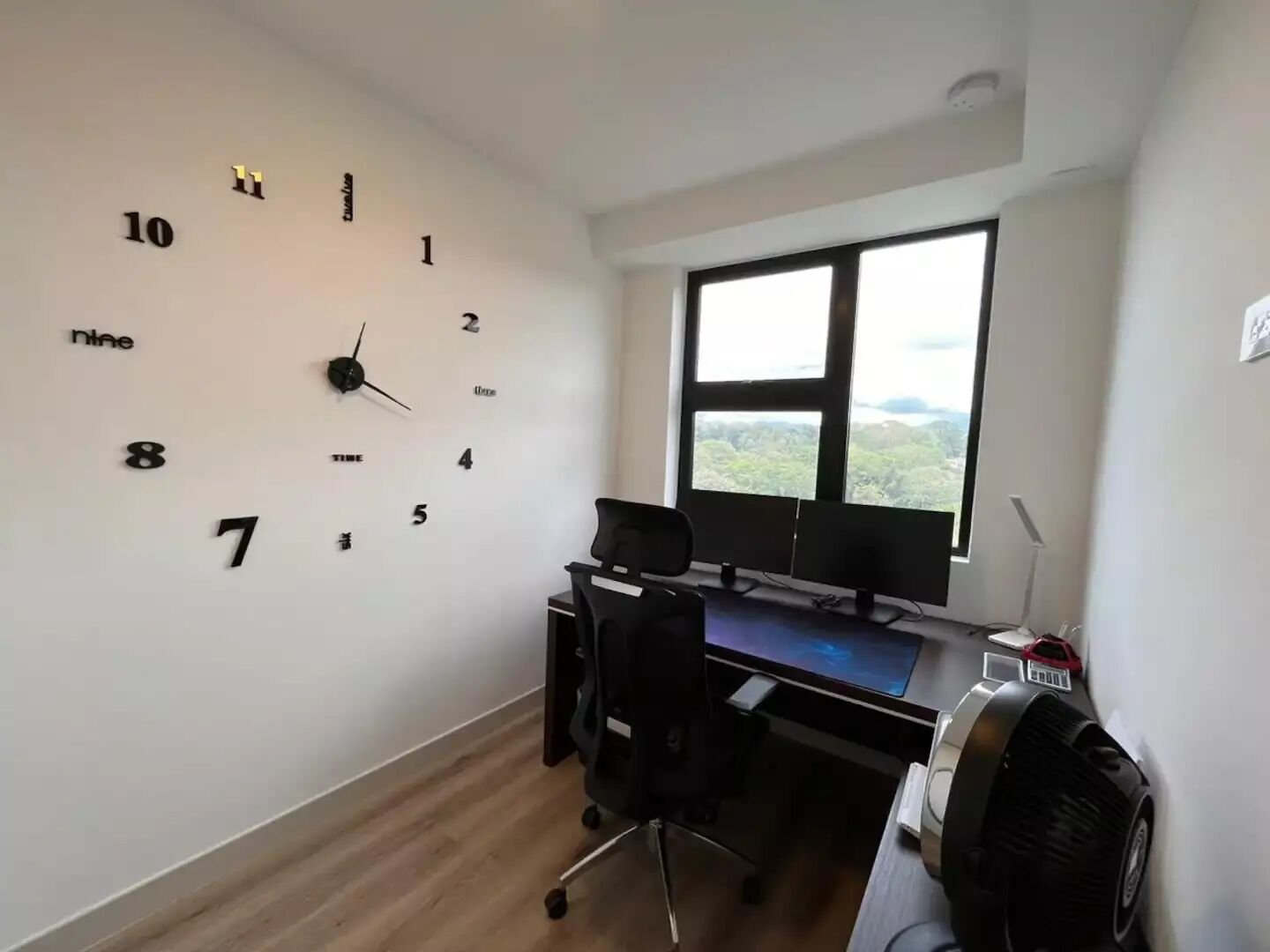A room with a wall clock, a desk with a computer, and a window with a view of greenery.