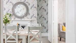 Un coin repas avec une table blanche dressée pour quatre personnes, un miroir décoratif au-dessus de la cheminée et un papier peint à motifs représentant des paons.