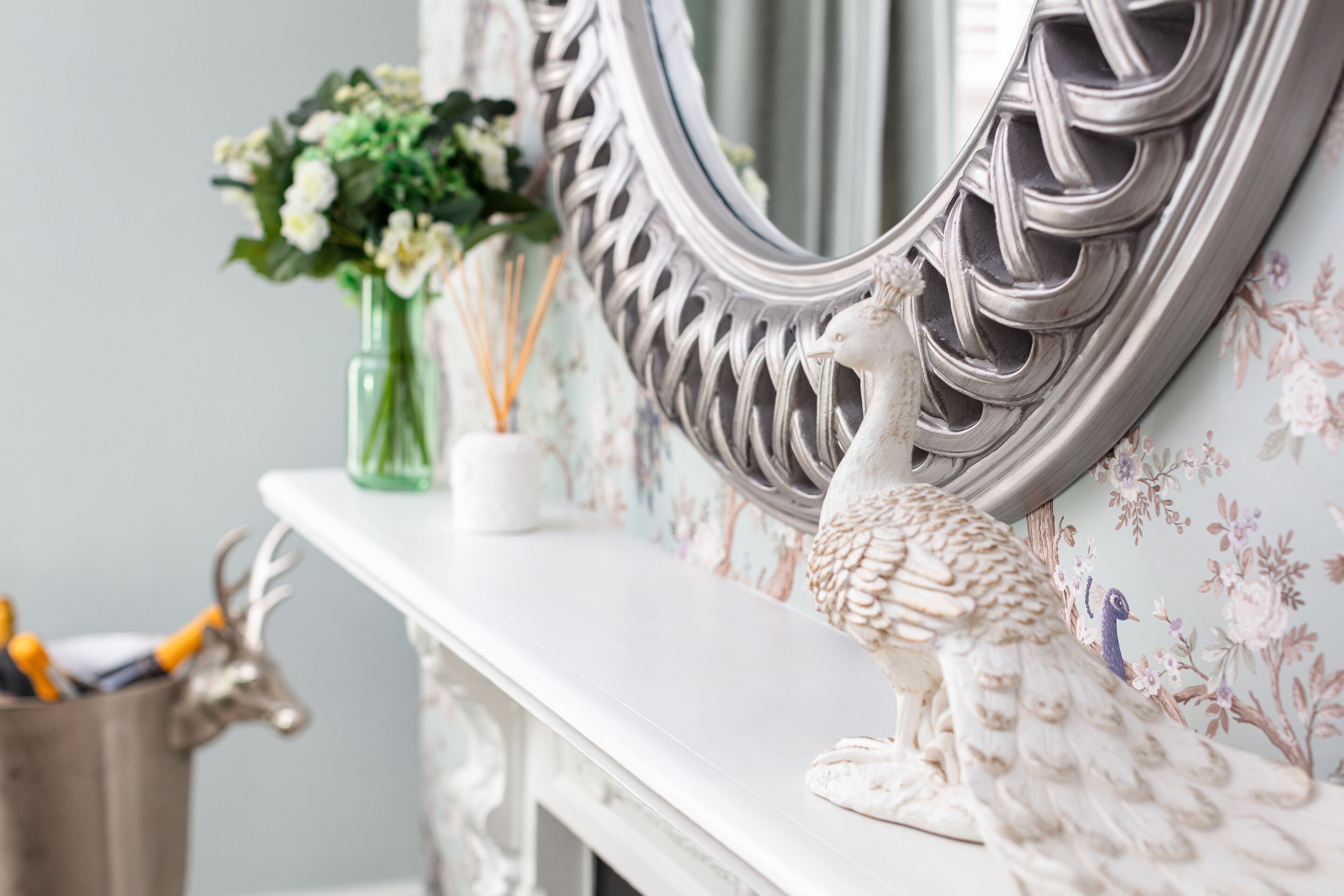 A decorative mantelpiece with a white ceramic peacock, a green vase with flowers, and a white candle.