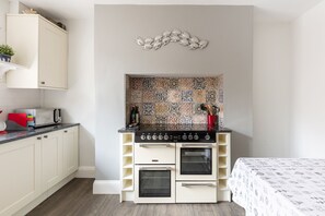 A compact kitchen with a stove, oven, and microwave. There is a patterned backsplash and a bed with a patterned bedspread.
