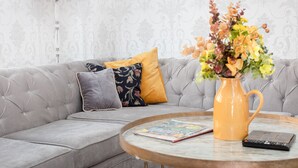 Un canapé gris agrémenté de plusieurs coussins, une table basse ronde sur laquelle reposent un livre et une télécommande, et un vase rempli de fleurs.