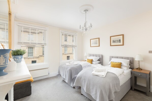 A hotel room with two beds, a bedside table, a lamp, and a window with blinds.