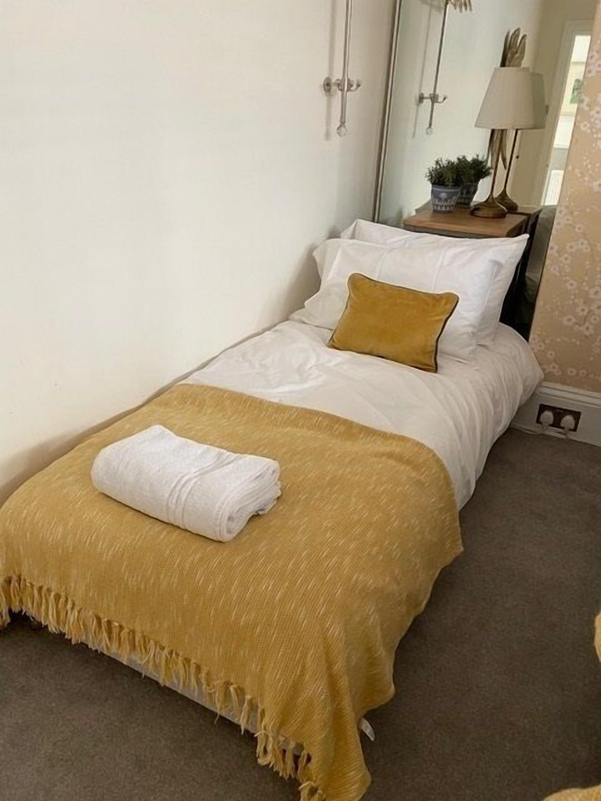 Un lit simple avec une couvre-lit blanche et jaune moutarde, une serviette blanche et une petite table de chevet avec une lampe et une plante.