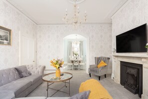 A living room with a sofa, armchair, and a fireplace. There is a television mounted on the wall and a chandelier hanging from the ceiling.