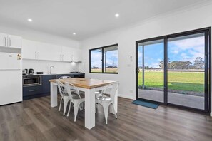 Une cuisine moderne avec des meubles blancs, une table à manger en bois et des chaises, ainsi qu’une grande porte-fenêtre coulissante donnant sur un espace extérieur verdoyant.
