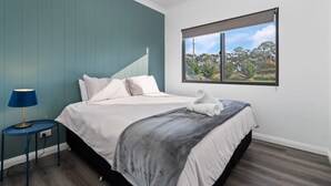 Une chambre à coucher avec un lit, une table de chevet avec une lampe, une fenêtre donnant sur des arbres et un mur bleu.