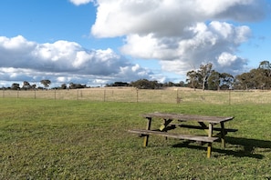 A picnic table in a grassy field with a fence in the background and trees.