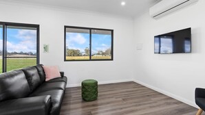 Une pièce moderne avec un canapé en cuir noir, un pouf vert, un téléviseur à écran plat fixé au mur et de grandes fenêtres offrant une vue sur l’extérieur.