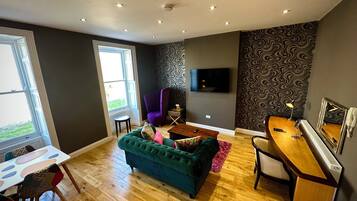 A modern living room with a green sofa, a wooden desk, a purple chair, and a TV on the wall.