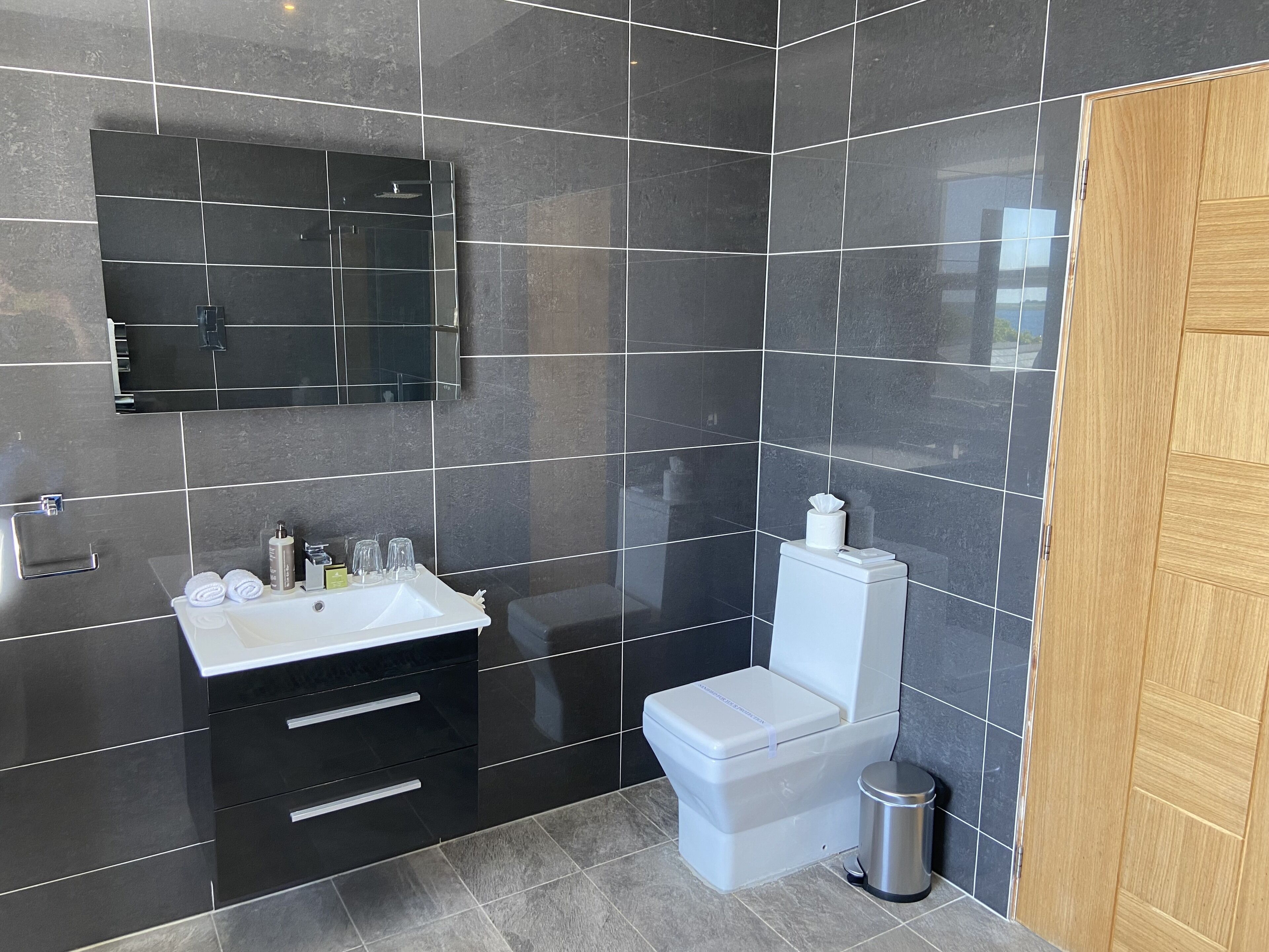 Un baño moderno con baldosas oscuras, un lavamanos blanco, un espejo y un inodoro.