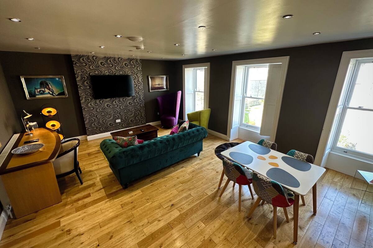 A modern living room with a teal sofa, a dining table with chairs, a wooden coffee table, a purple armchair, and a flat-screen TV mounted on the wall.