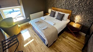 Une chambre avec un plancher en bois, un lit recouvert d’une couette à rayures blanches et grises, deux lampes de chevet, une chaise et une fenêtre donnant sur un parking.