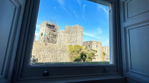 Une vue à travers la fenêtre d’un bâtiment en pierre, sur laquelle on aperçoit un drapeau, peut-être un site historique, ainsi que quelques plantes.