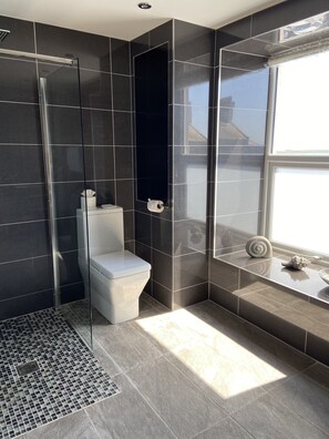A modern bathroom with dark tiles, a glass shower enclosure, a toilet, and a window with a view. - Halvard Apartments at Castletown (Castletown)