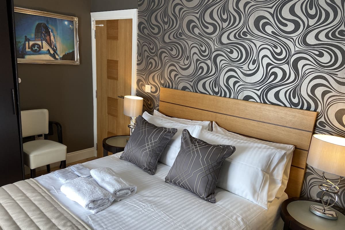 A neatly made bed with pillows and a folded towel, a wooden headboard, a patterned wall, a small table with a lamp, and a chair.