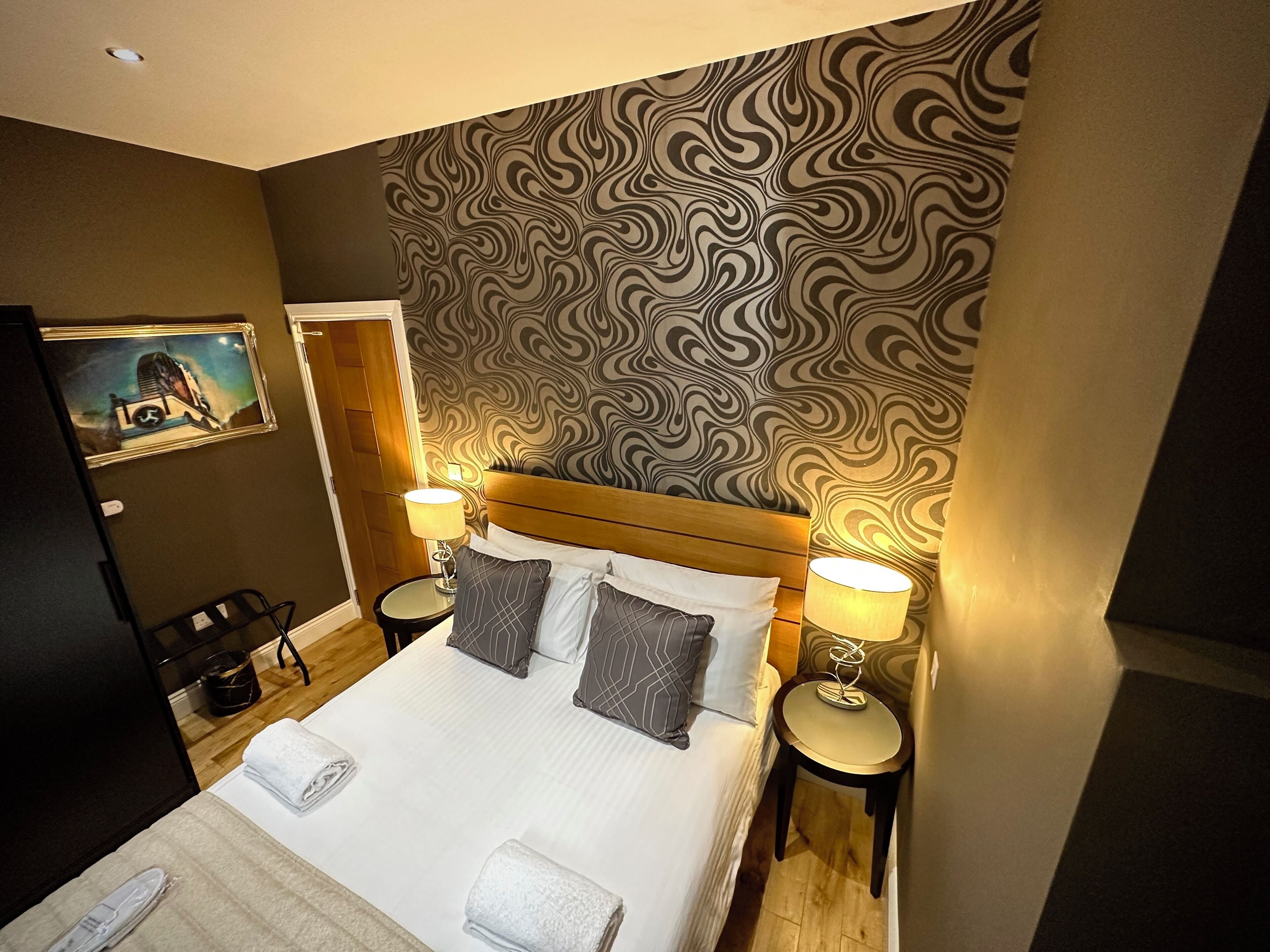 Une chambre d’hôtel avec un grand lit, deux tables de chevet avec des lampes, une tête de lit en bois, un papier peint à motifs et un tableau au mur.
