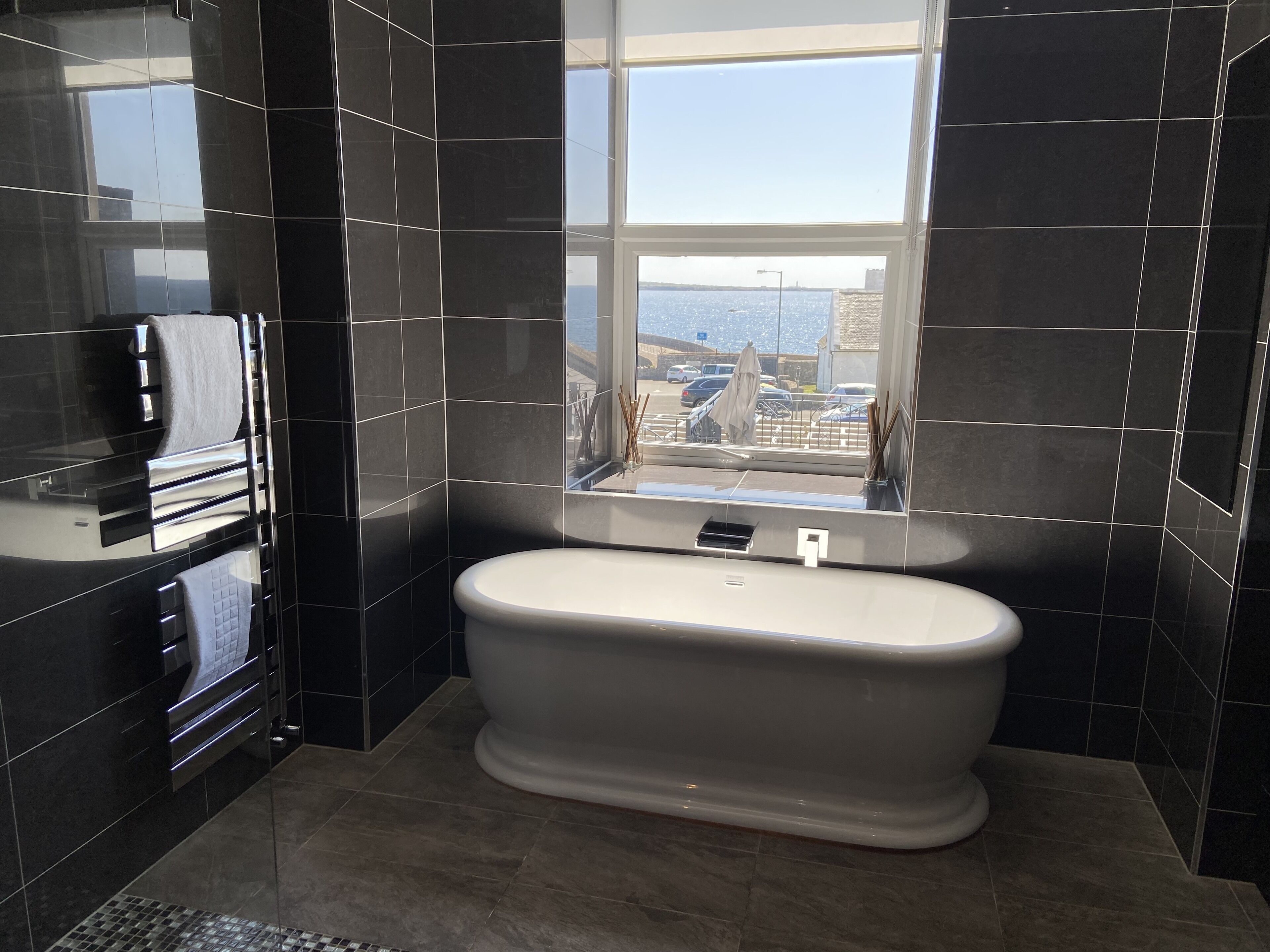 Un baño moderno con una bañera independiente, un amplio ventanal con vistas a la ciudad y paredes de azulejos oscuros.