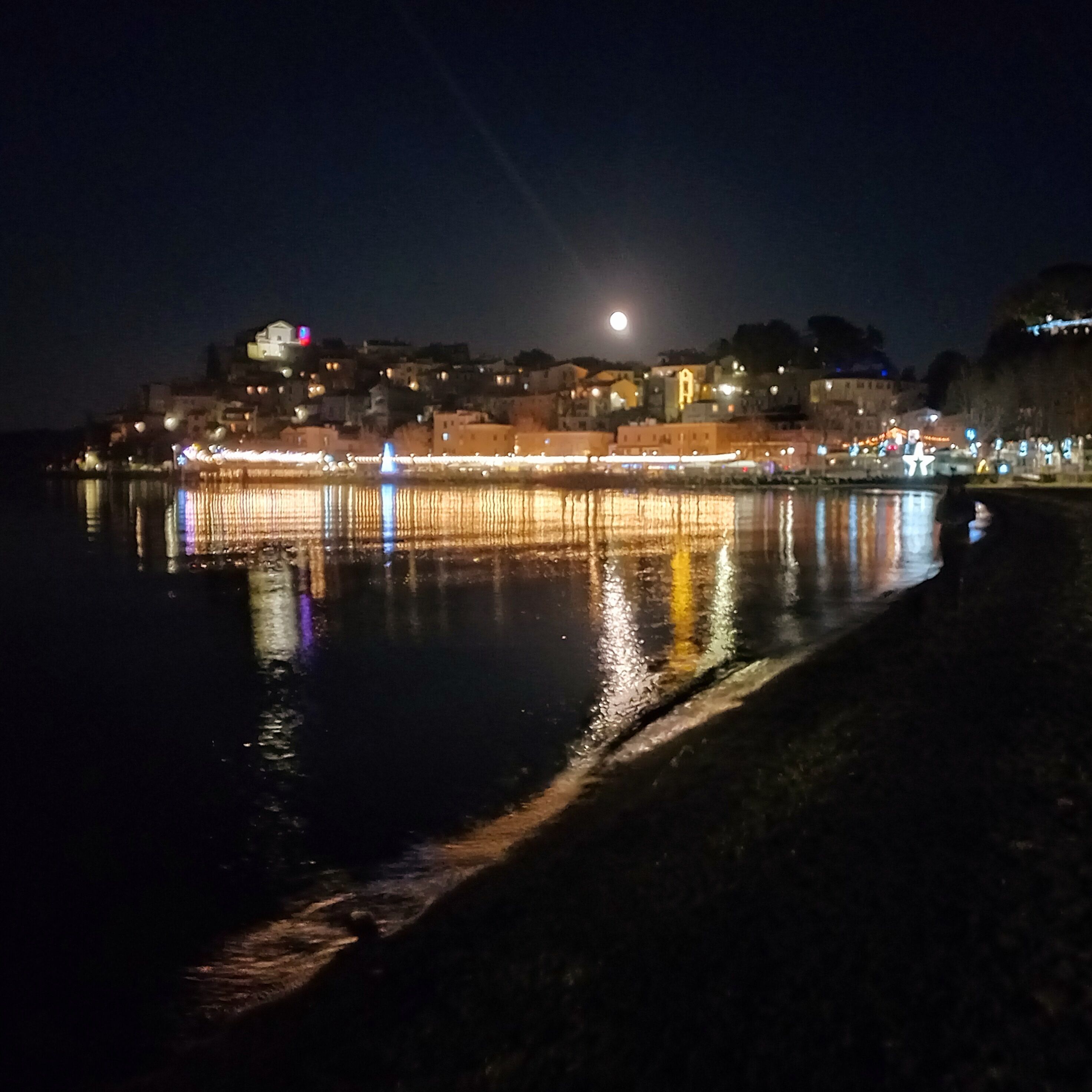 A nighttime coastal scene with a calm sea reflecting city lights and a prominent moon in the sky.