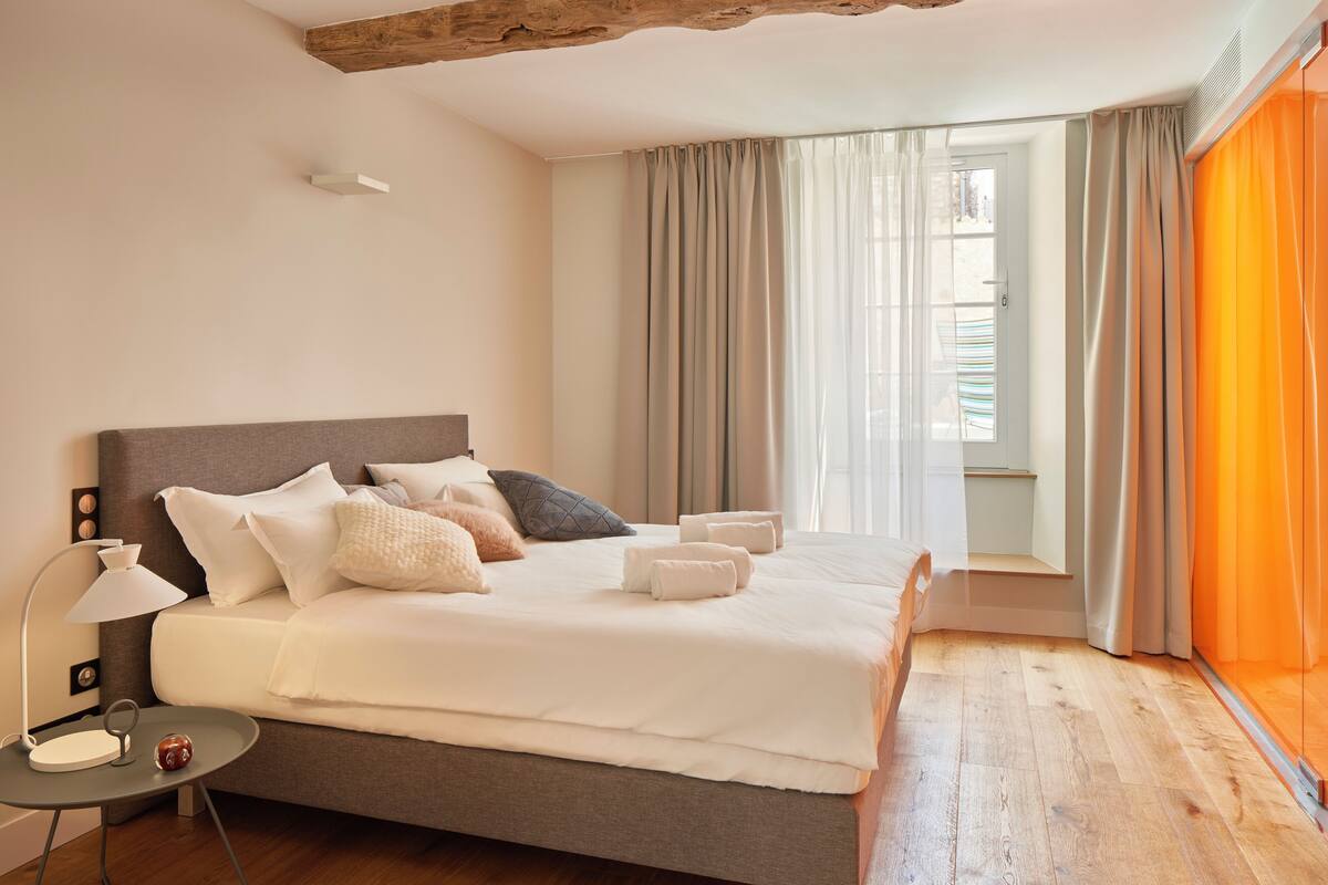 A modern bedroom with a large bed, a bedside table with a lamp, and a view of the outdoors through sheer curtains.