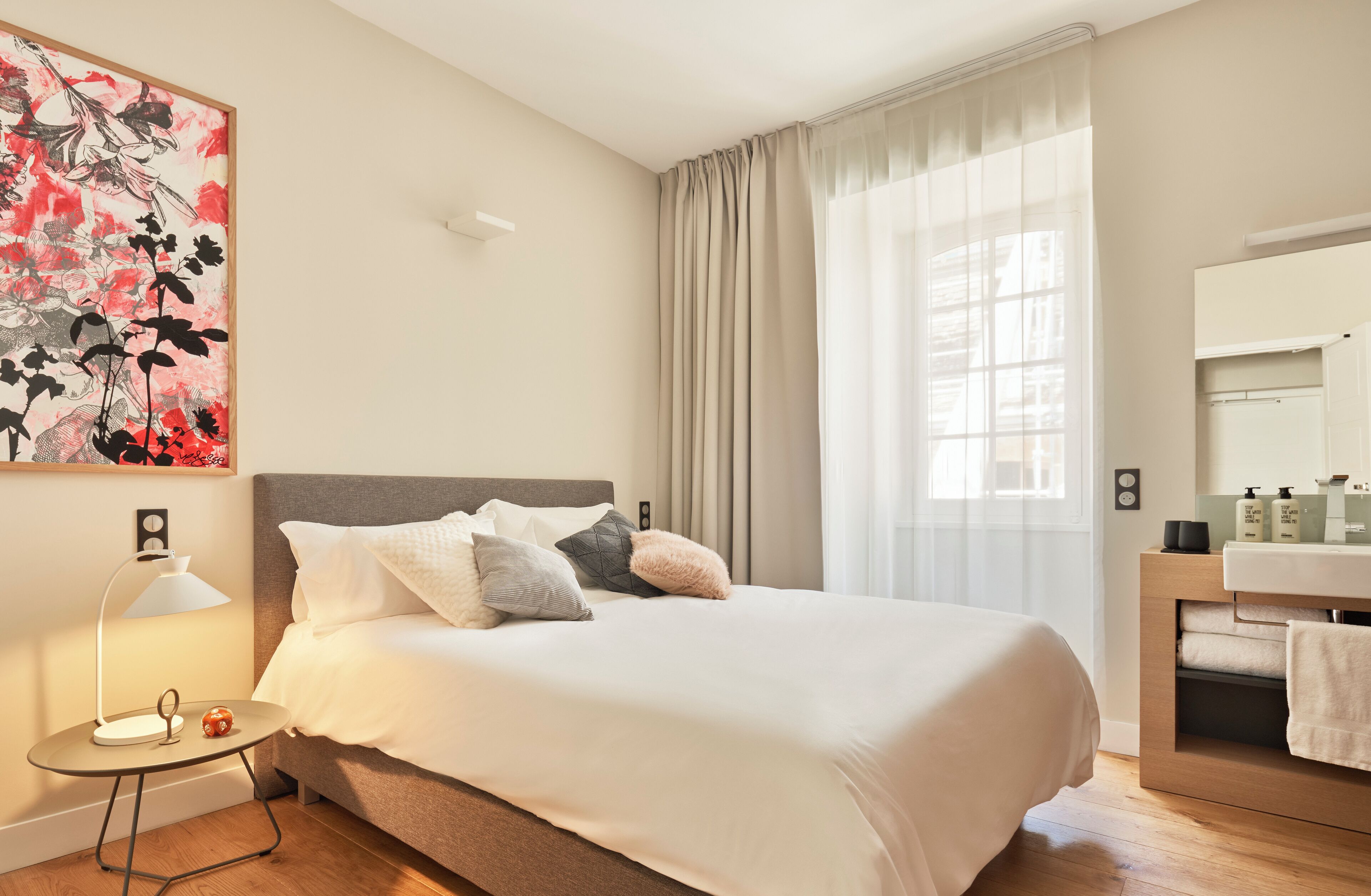 A well-lit bedroom with a large bed, a bedside table with a lamp, a nightstand, a mirror, and a window with curtains.