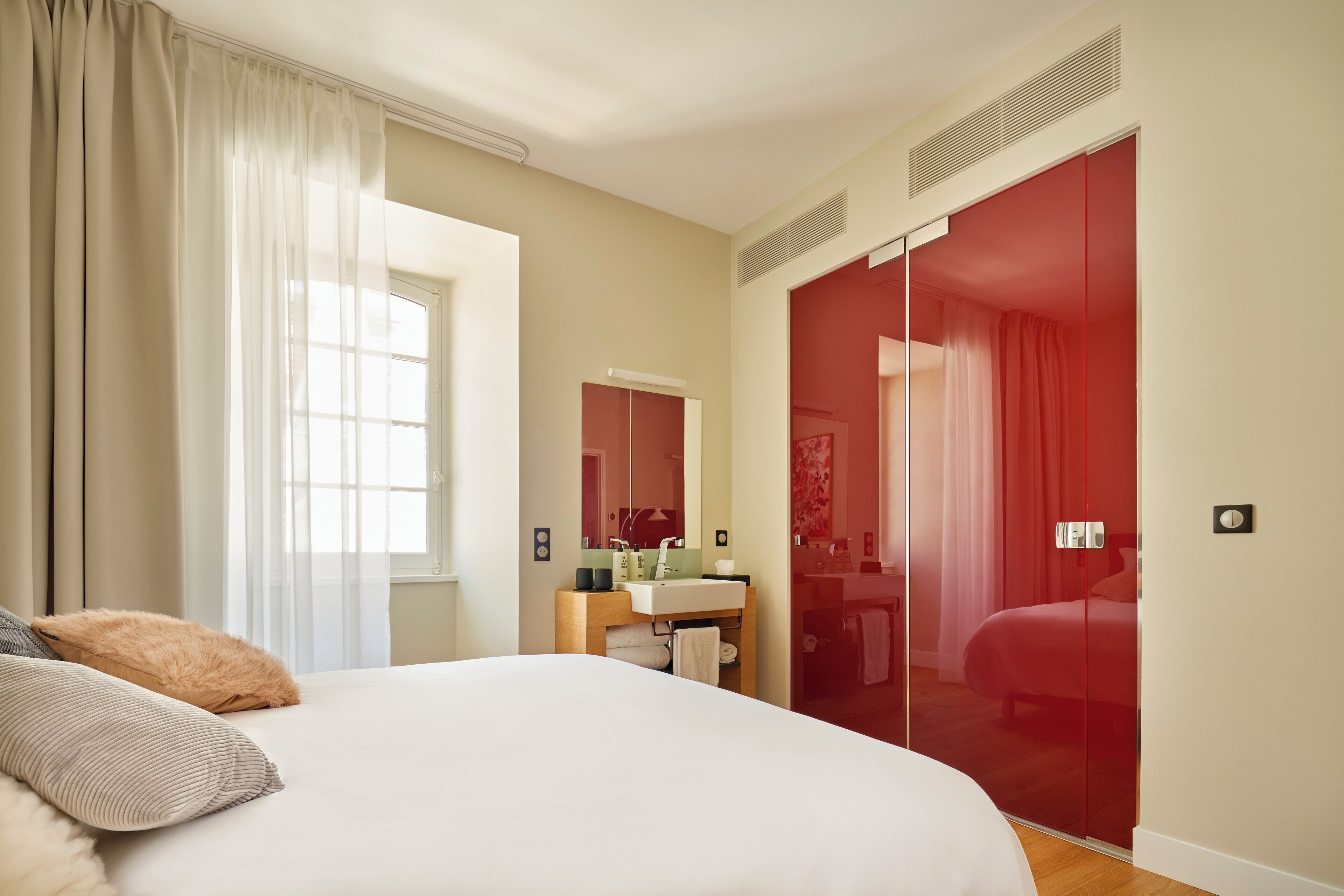 A bedroom with a large bed, a bedside table, a mirror, a red wardrobe, and a window with curtains.