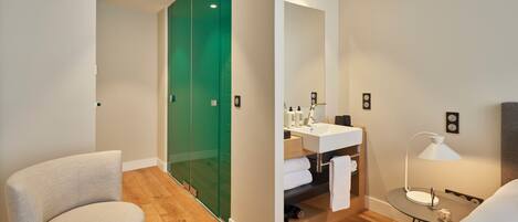 A modern bathroom with a glass shower enclosure, a sink with a mirror, and a bedside table with a lamp.