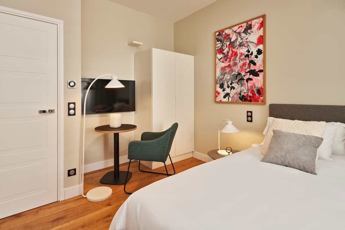 A hotel room with a bed, a desk with a chair, a TV, and a painting on the wall.