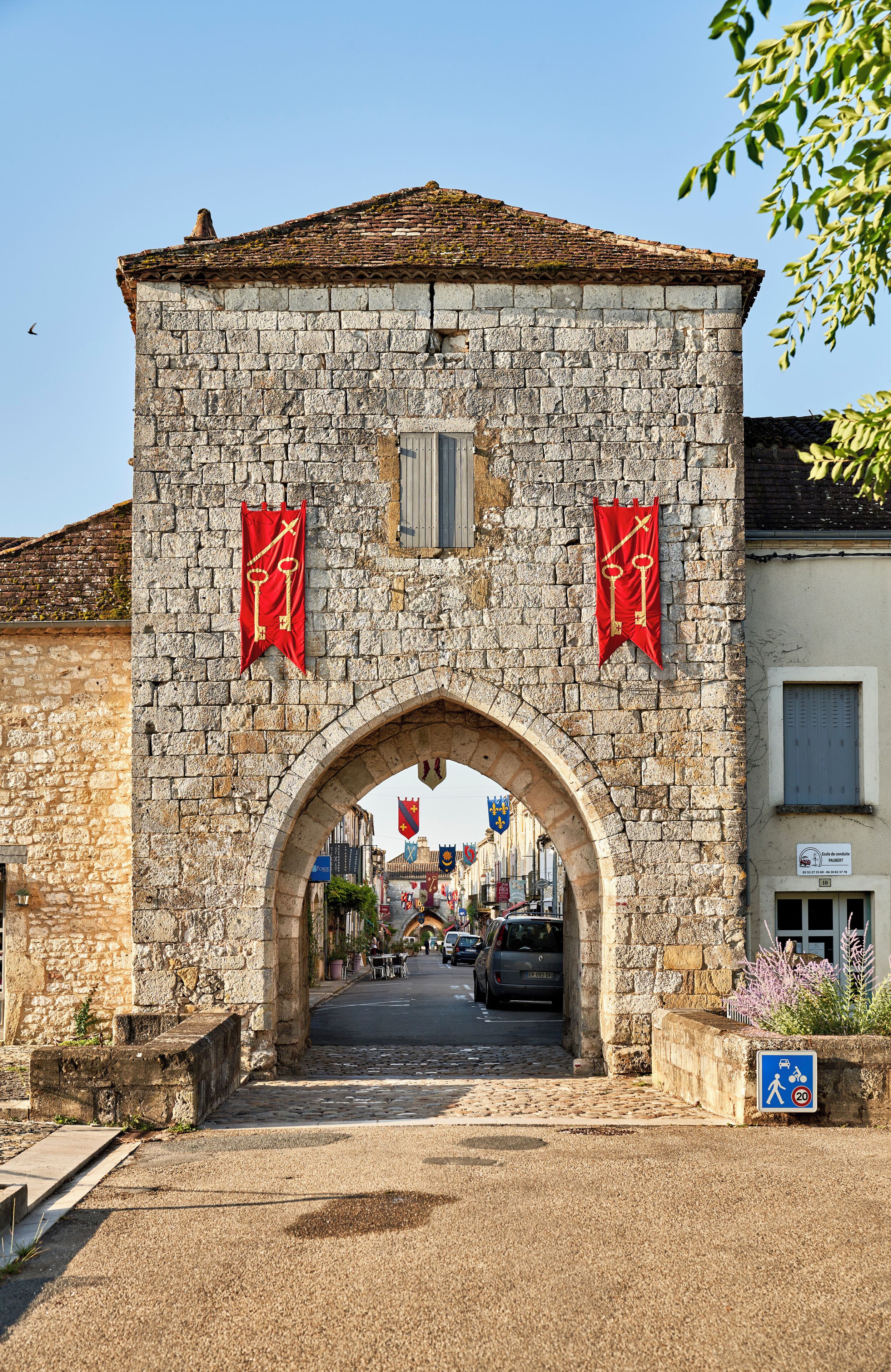 A stone archway with red banners, leading to a street with parked cars and buildings.