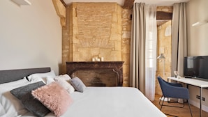 A bedroom with a bed, a desk, a chair, a stone wall, and a fireplace. - Hotel Le Chevalier Bleu (Monpazier)