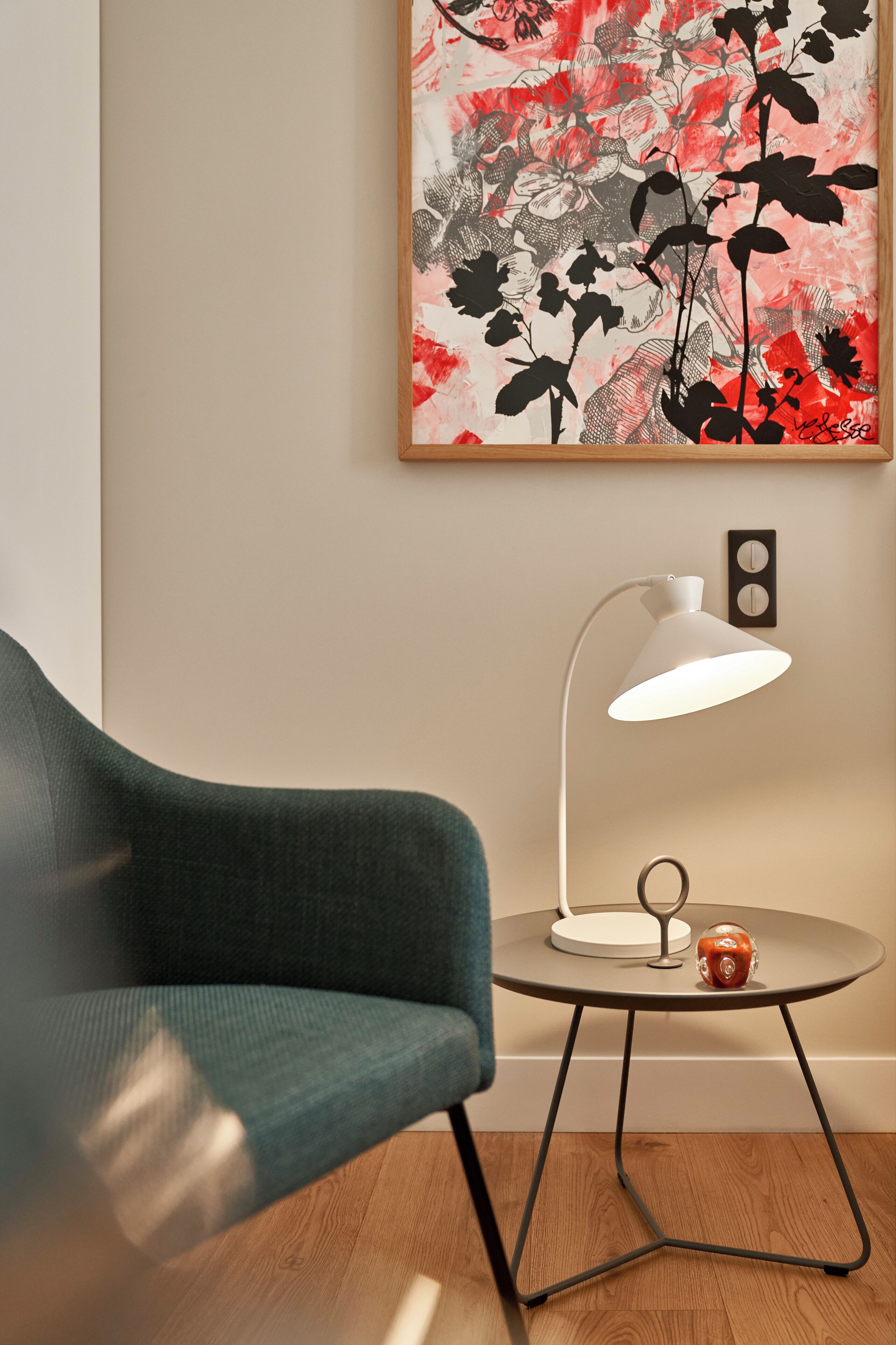 A modern room with a green armchair, a round table with a lamp, and a floral artwork on the wall.