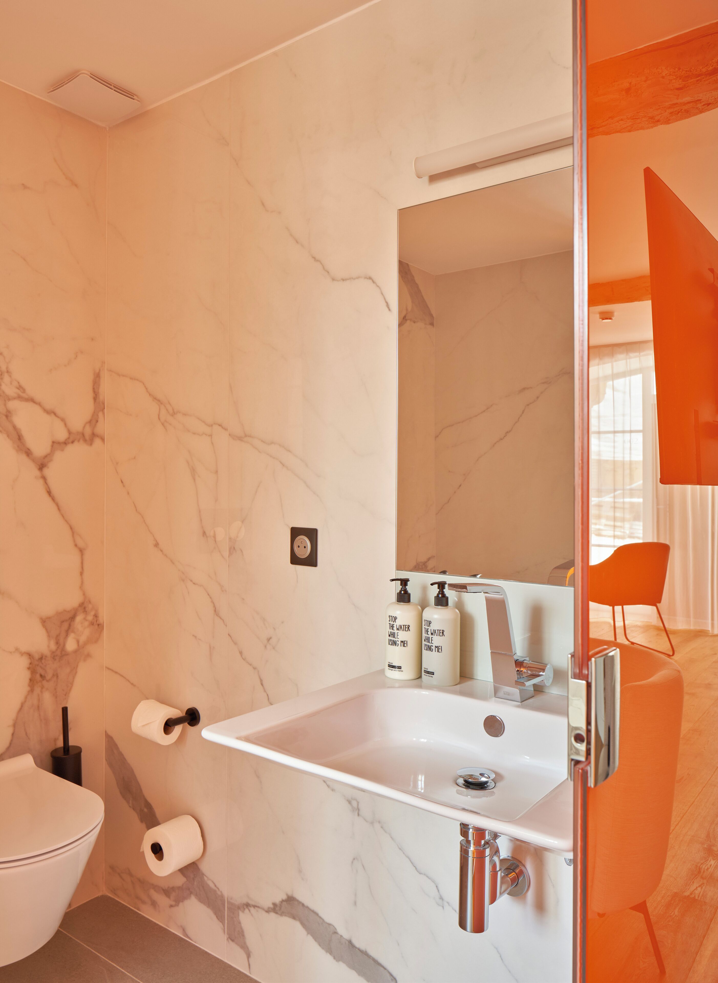 Un baño moderno con pared de mármol, un lavamanos blanco con un espejo arriba y dos botellas de jabón para manos.