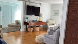 A spacious living room with a wooden floor, a television mounted on the wall, and a sofa. - Ocean View cottage in Indian Neck. Super Host with 5-Star Rating (Branford)