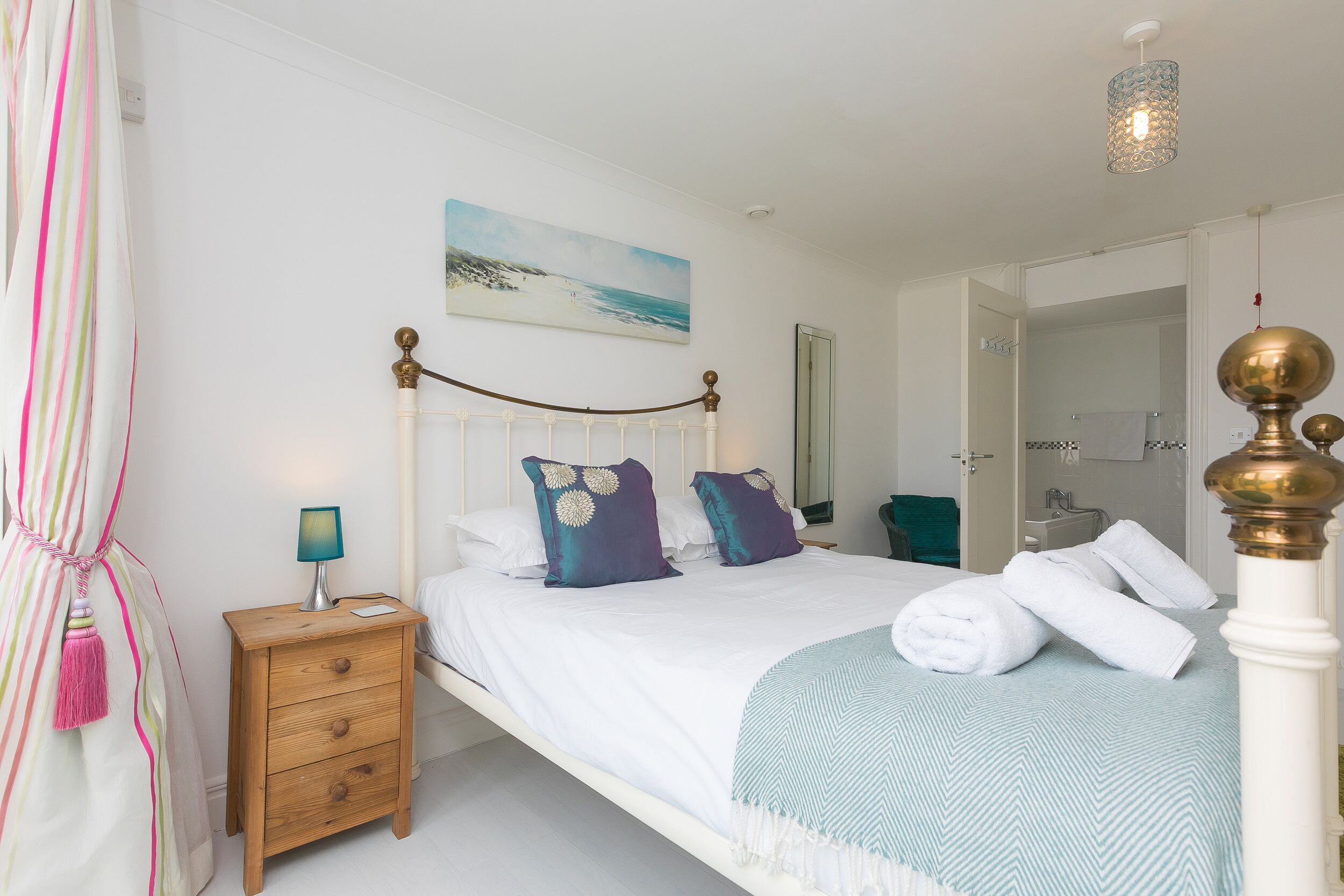 A bedroom with a bed, a nightstand, a lamp, a painting, and a bathroom visible through an open door.