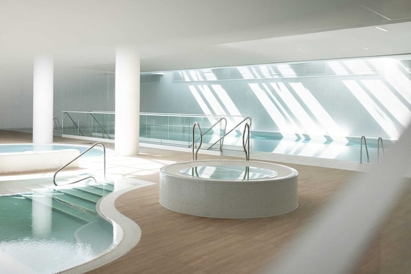 A modern indoor swimming pool with a circular hot tub, clear glass railings, and a view of the sky through the ceiling.
